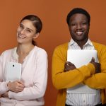 Stylish young mixed race team of two coworkers using electronic gadgets for work, positive cheerful white female carrying digital tablet and handsome smiling black male embracing laptop computer